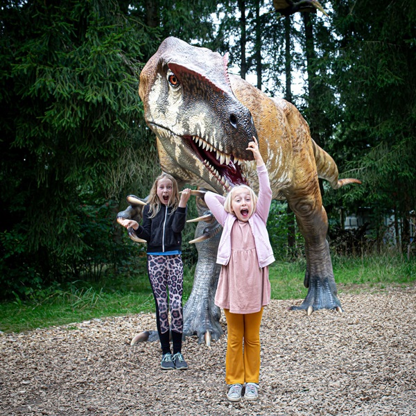 Two girls jump excitedly in front of a dinosaur in the dinosaur park at GIVSKUD ZOO – ZOOTOPIA